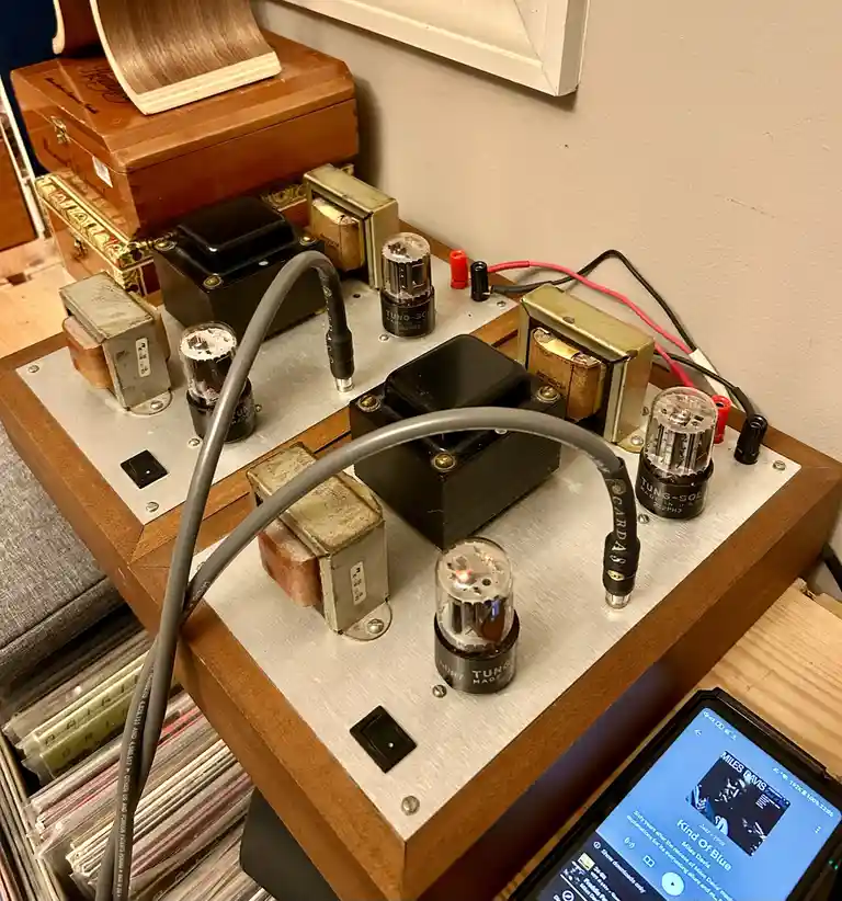 A close-up photo shows a vintage tube amplifier with glowing vacuum tubes and transformers, connected to a smartphone…