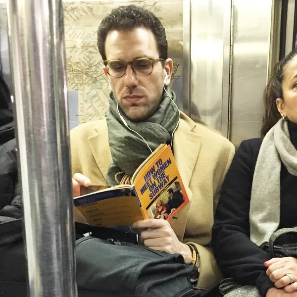 A man wearing glasses and a scarf sits on a subway train, holding and reading a book titled 'How to Meet Women on the…