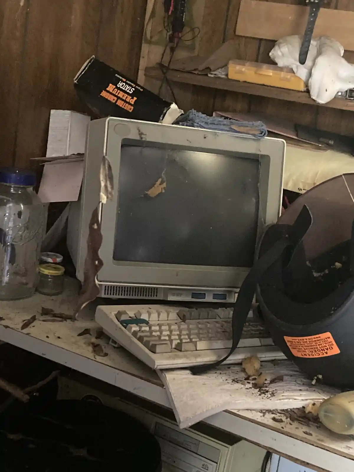 An old, dusty CRT computer monitor and keyboard sit on a cluttered, dirty shelf surrounded by various items including a…