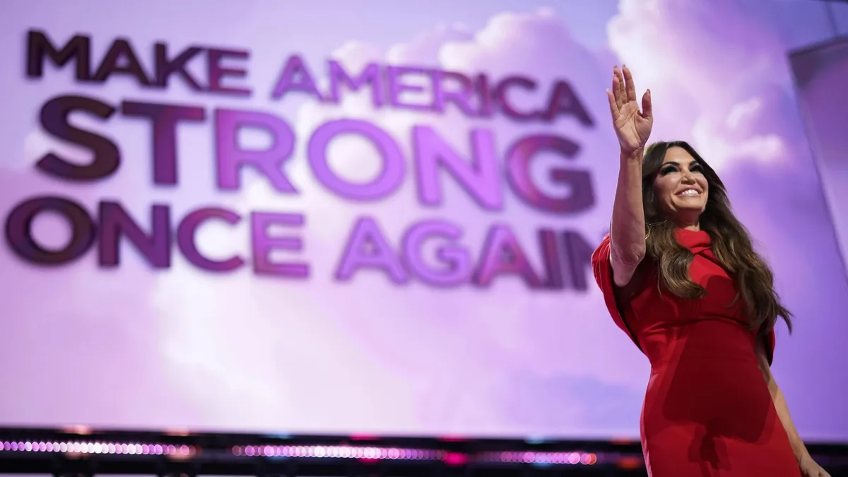 A woman in a red dress waves to an audience while standing in front of a large screen displaying the slogan 'MAKE AMERICA…