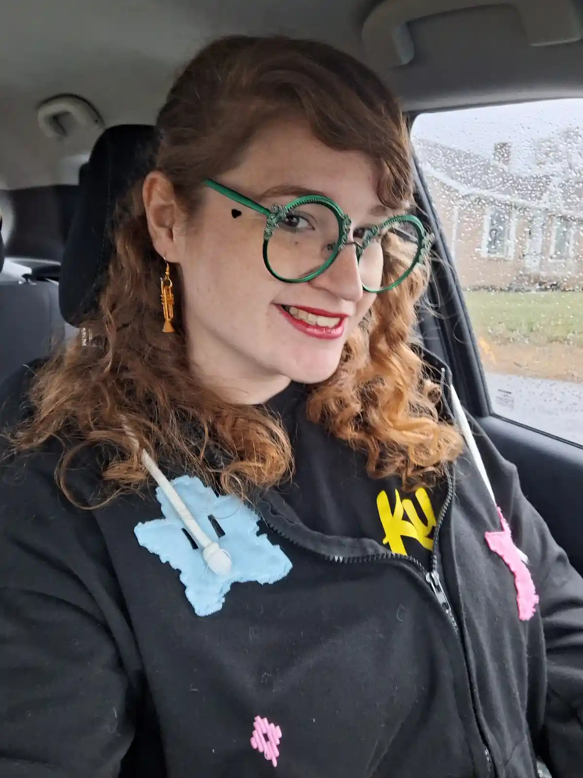 A person with curly reddish-brown hair and green round glasses sits in a car with raindrops on the window, smiling. They…