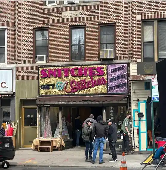A storefront with a large, glittery sign that reads 'SNITCHES GET STITCHES' in pink and black lettering, with a needle and…