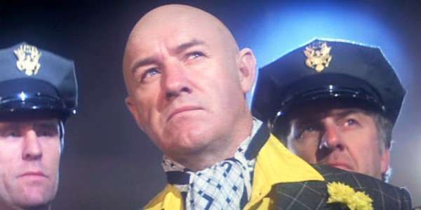 A still from a movie shows a bald man in a yellow jacket and checkered bowtie looking upward with a skeptical expression…