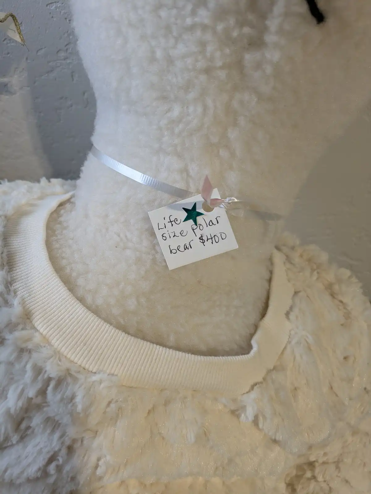 A close-up of a large, fluffy, cream-colored stuffed bear wearing a cream sweater with a ribbed collar. A handwritten tag…