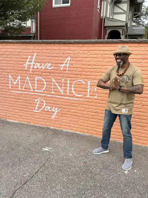 A smiling man in a straw hat and sunglasses stands in front of a peach-colored brick wall with the phrase 'Have A MADNICE…