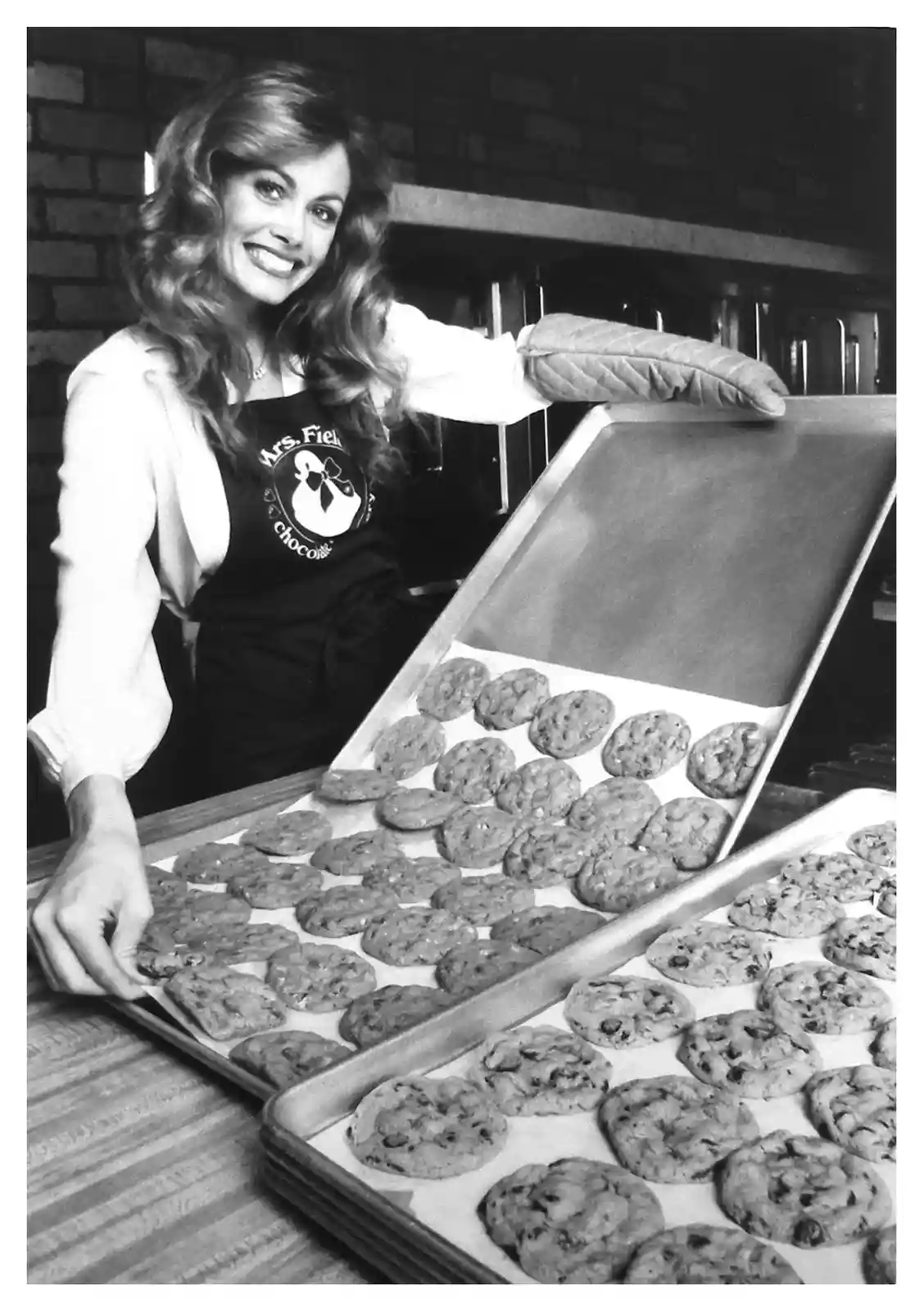 A black-and-white photo shows a smiling woman with long hair wearing a 'Mrs. Field's chocolate' apron, holding up a baking…