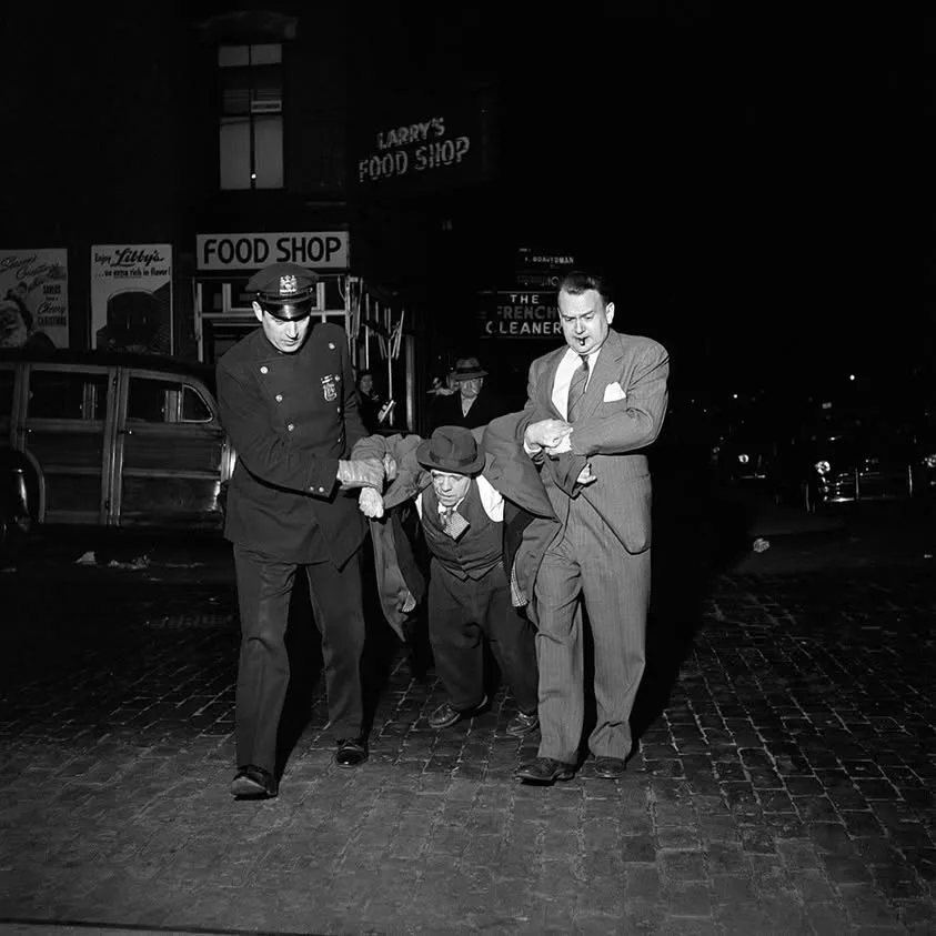 A black-and-white photo from the 1940s shows a police officer and a man in a suit dragging a small man in a hat and vest…