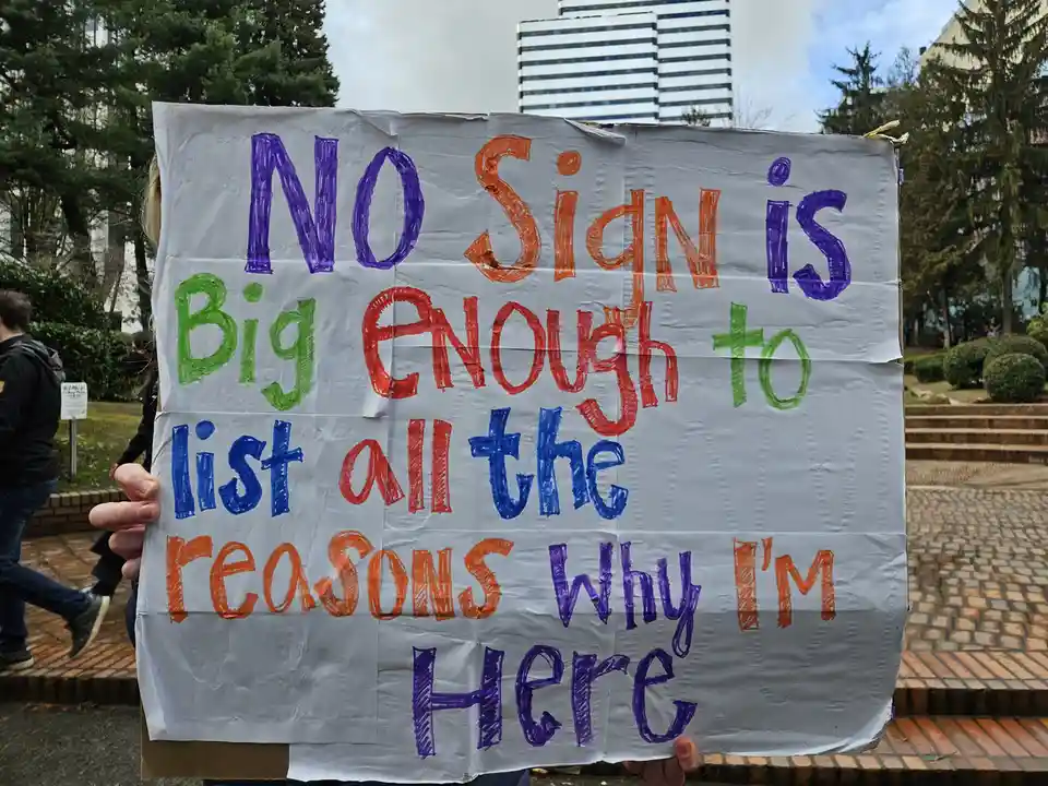 A person holds a handmade protest sign with colorful, childlike marker writing that reads 'No Sign is Big Enough to list all…