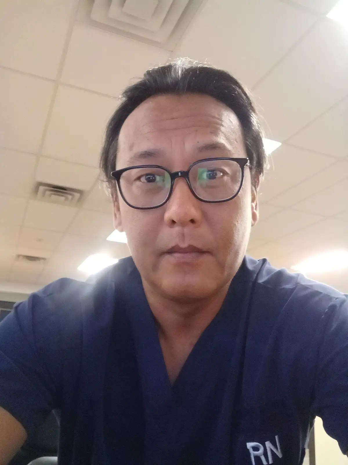 A selfie of a middle-aged man with dark hair and black-rimmed glasses, wearing blue medical scrubs with 'RN' embroidered on…