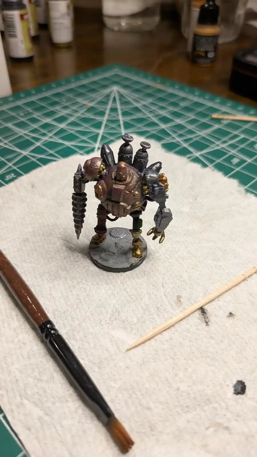 A painted miniature figure, resembling a heavily armored, multi-limbed creature with purple and gold accents, stands on a…