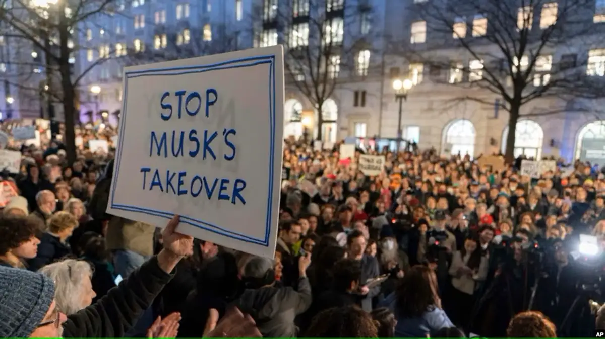 A large nighttime protest crowd holds signs, with one prominent white sign in the foreground reading 'STOP MUSK'S TAKEOVER'…
