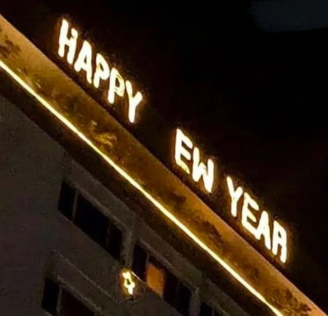 A nighttime photo of a building's illuminated sign that reads 'HAPPY EW YEAR', with 'EW' replacing 'NEW' due to a blurry or…