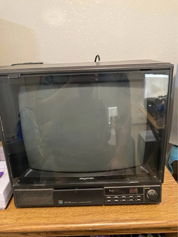 A black, boxy, vintage Daytron CRT television sits on a wooden surface, its screen dark and reflective, showing a faint…