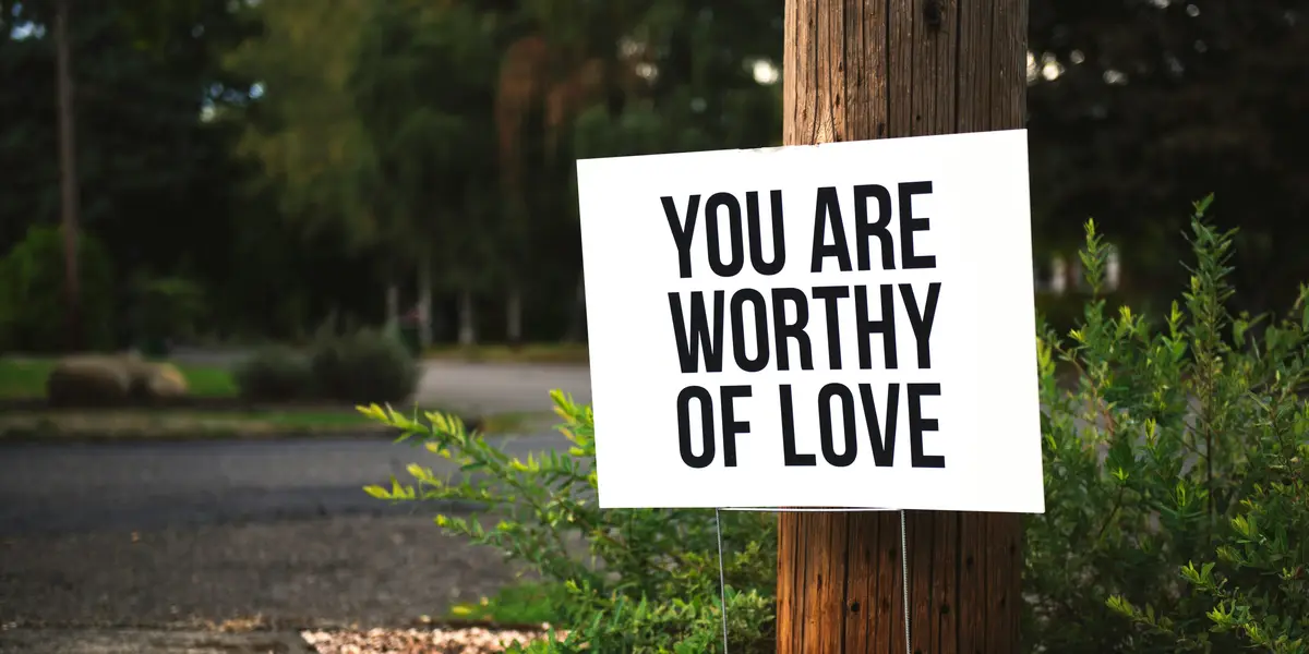 A white sign with bold black text reading 'YOU ARE WORTHY OF LOVE' is taped to a wooden utility pole beside green bushes and…