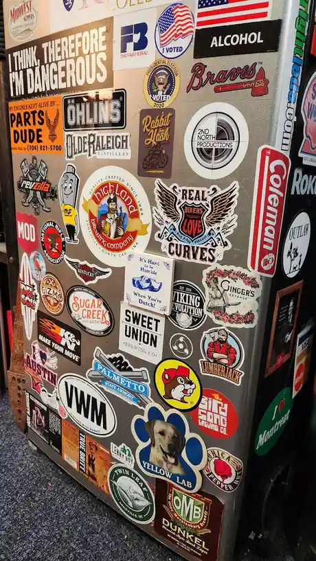 A close-up of a refrigerator covered in numerous stickers, including brands, political slogans, and humorous phrases…