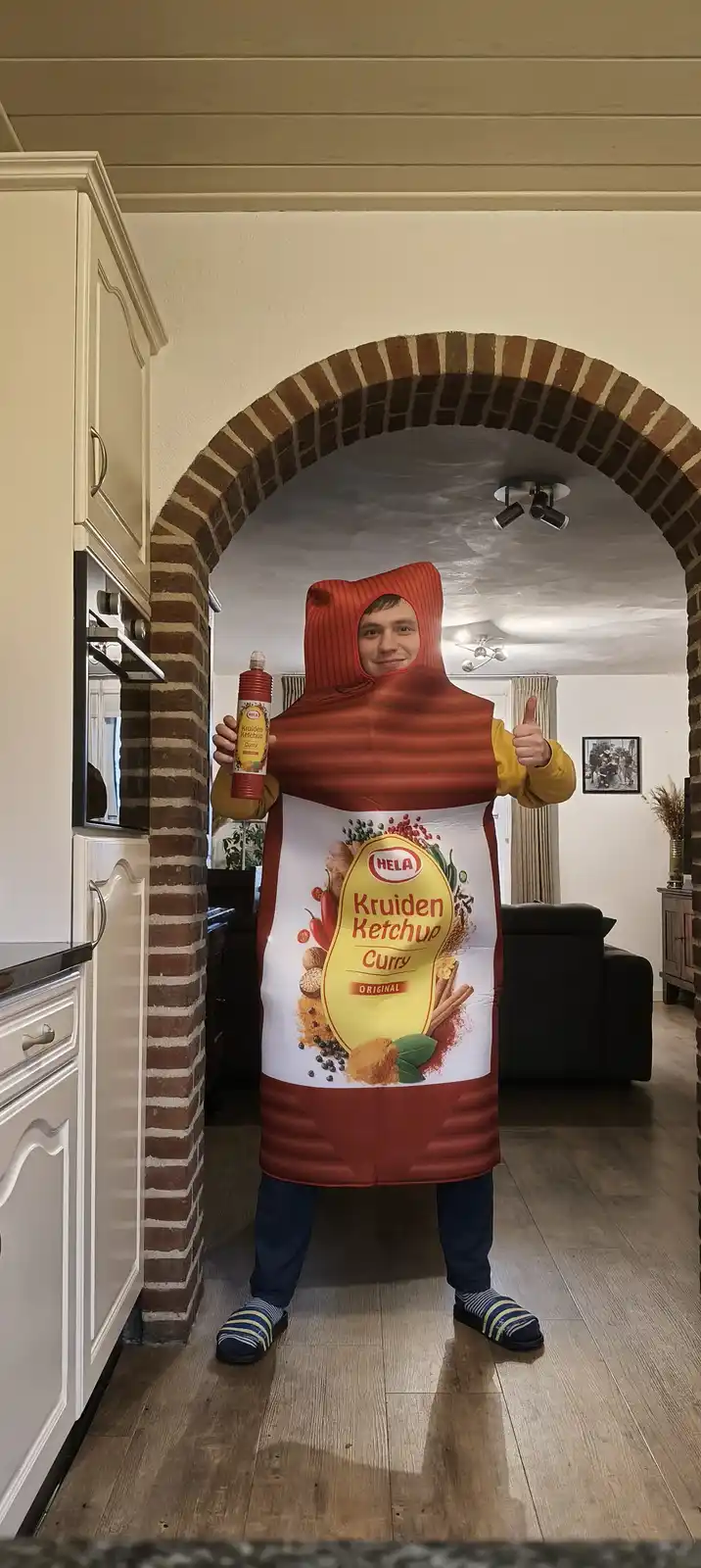 A person stands in a kitchen doorway wearing a full-body costume resembling a red bottle of Hela Kruiden Ketchup Curry…