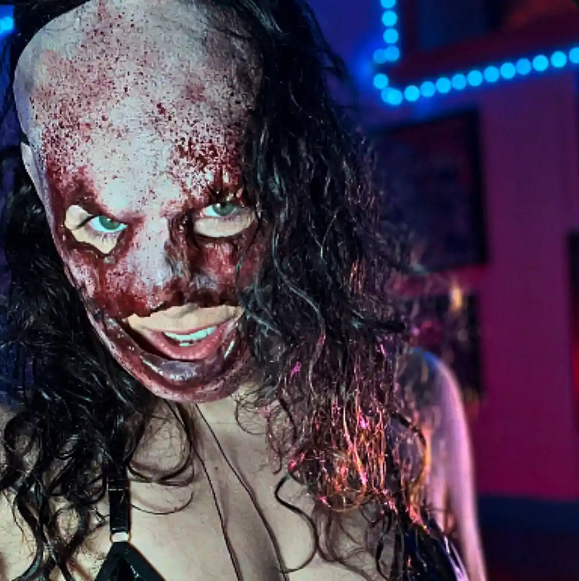 A person with dark curly hair and dramatic zombie makeup featuring smeared red blood effects, pale skin, and glowing green…