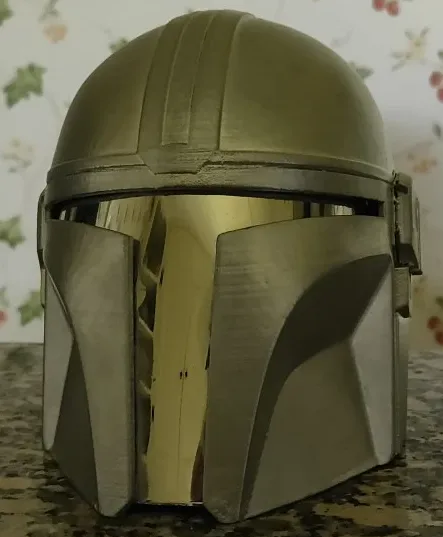 A close-up photo of a metallic Mandalorian helmet, likely a 3D-printed replica, resting on a speckled countertop. The…