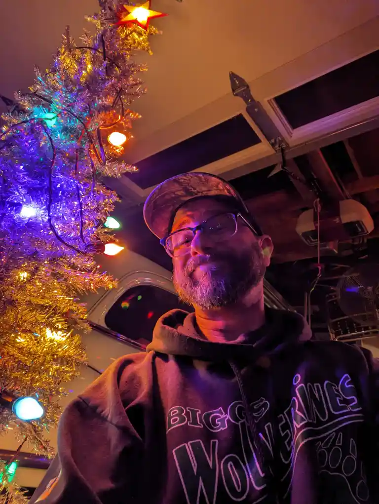 A man with glasses and a beard, wearing a camouflage cap and a 'Biggs Wolverines' hoodie, poses for a selfie in a dimly lit…