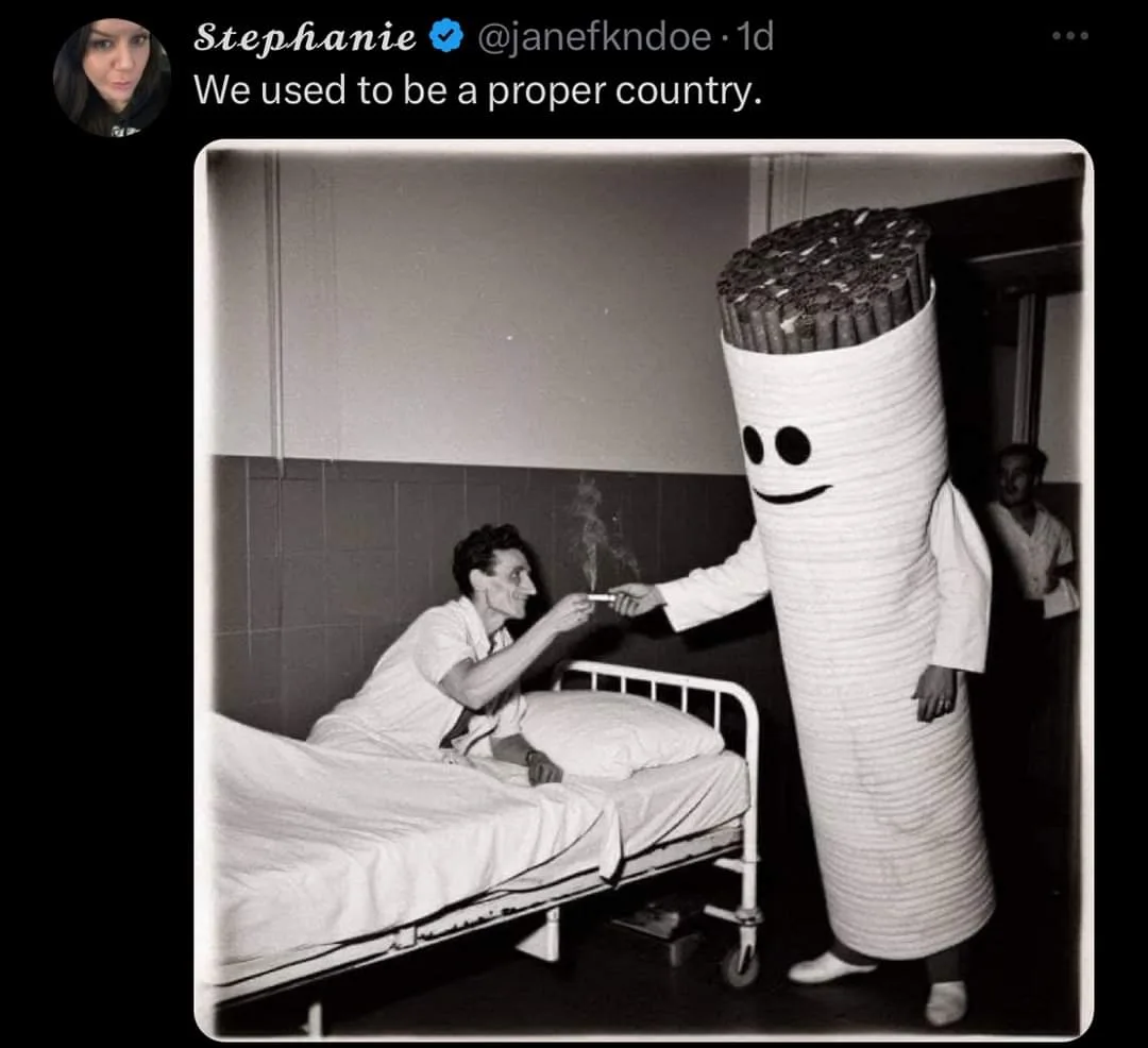 A black-and-white photo shows a man in a hospital bed smiling as a person in a large, smiling cigarette costume hands him a…