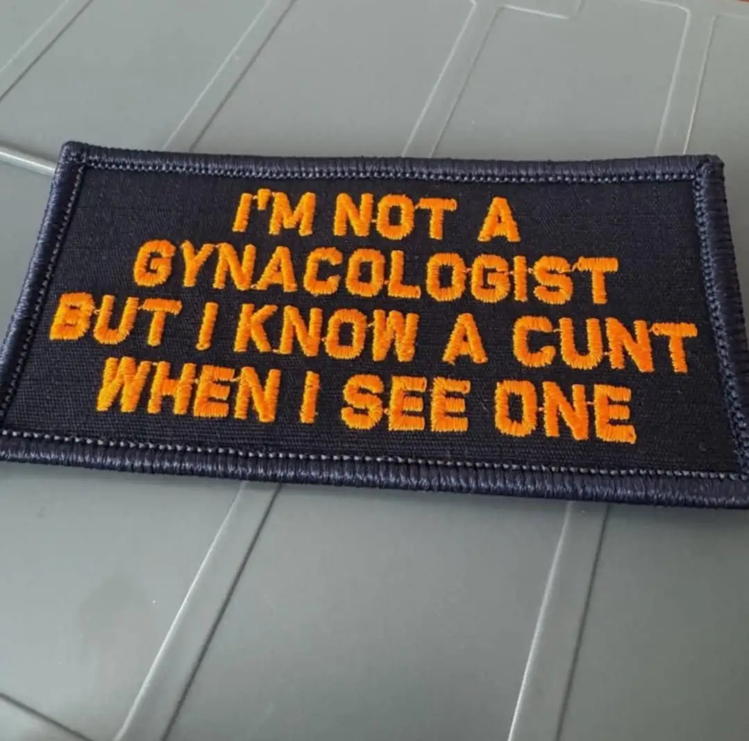 A dark blue rectangular embroidered patch with orange text reads 'I'M NOT A GYNACOLOGIST BUT I KNOW A CUNT WHEN I SEE ONE'…