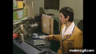 A woman in a yellow jacket sits at a radio station desk with a microphone and coffee cup. She initially looks at a plate of…