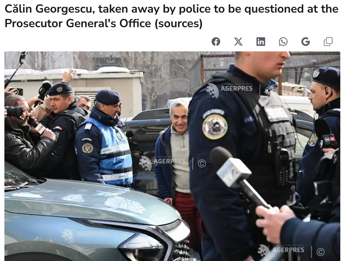 A man in a blue jacket and grey sweater smiles while being escorted by police officers near a car, surrounded by reporters…