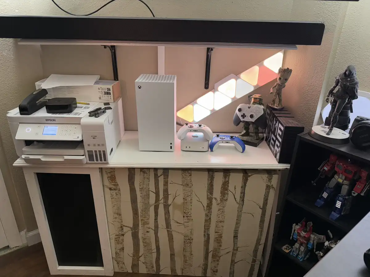 A white desk holds an Epson printer, an Xbox Series X console, and two game controllers, next to a glowing geometric light…