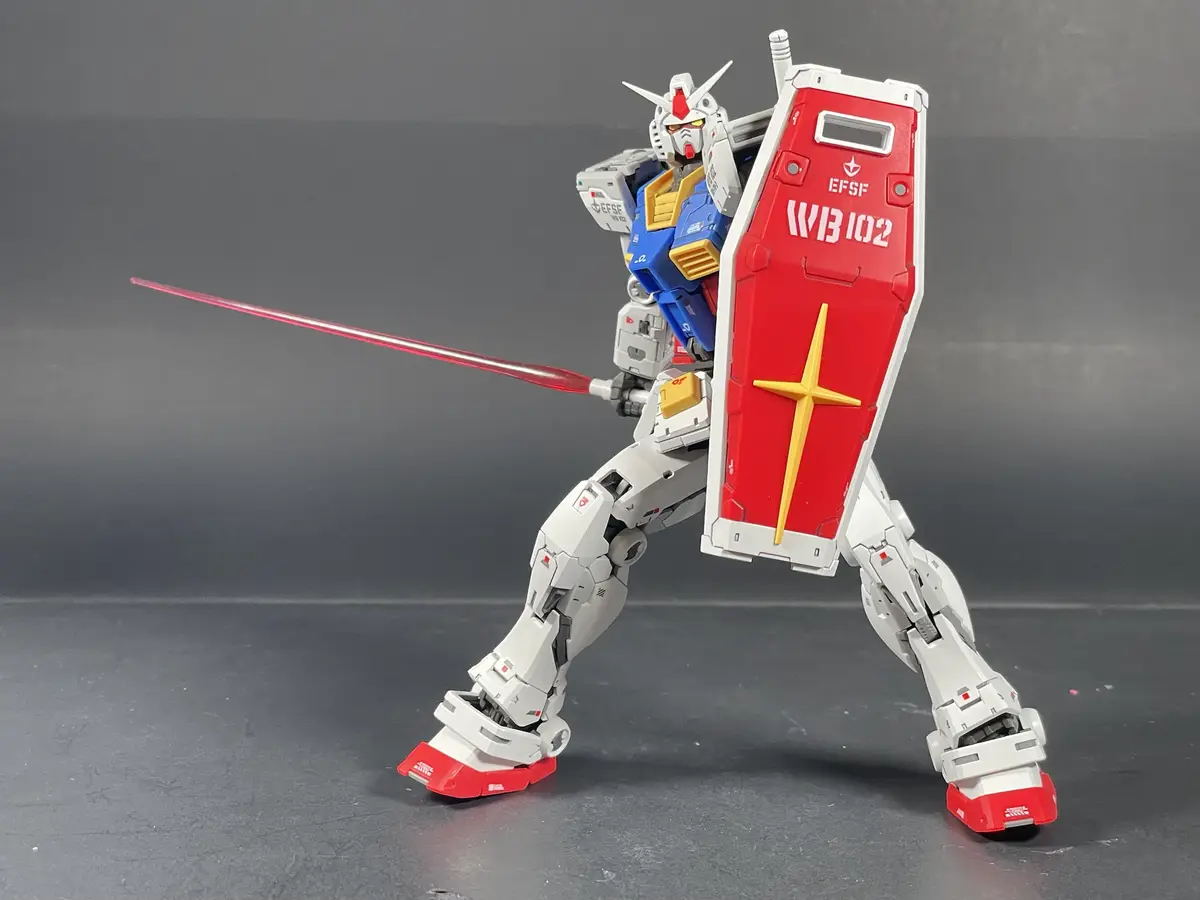 A white, blue, and red Gundam model stands in a dynamic pose holding a red shield with a yellow cross and a red beam saber…