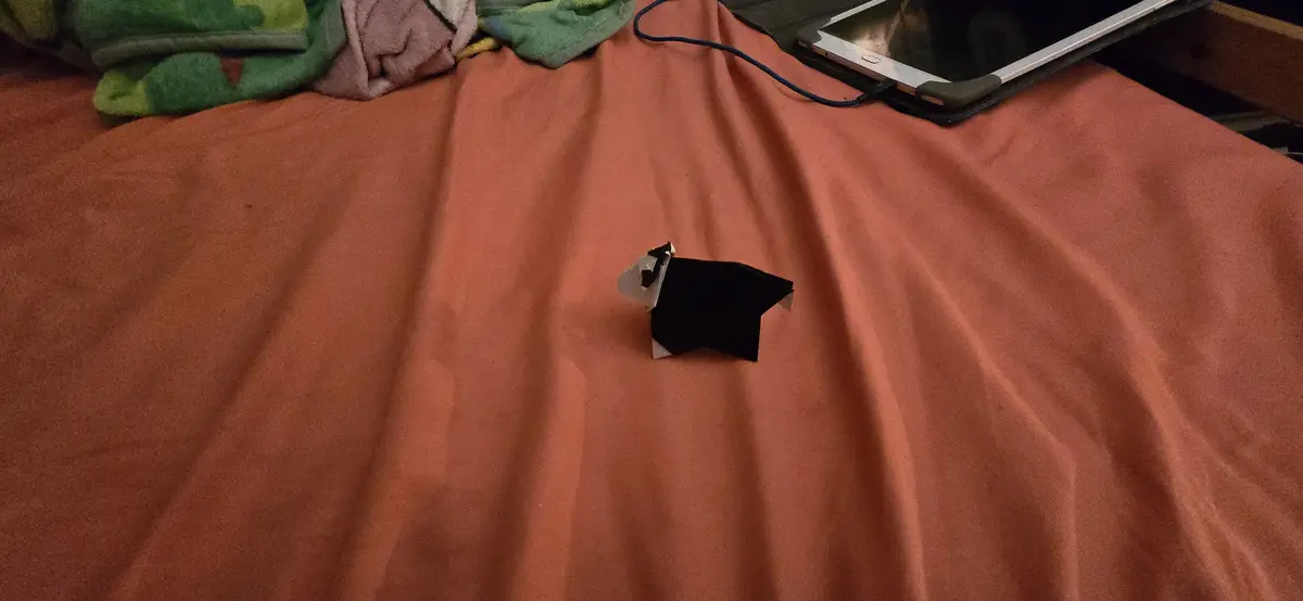 A small black-and-white origami dog sits alone on a rust-colored bedsheet, with a tablet and tangled cable in the…