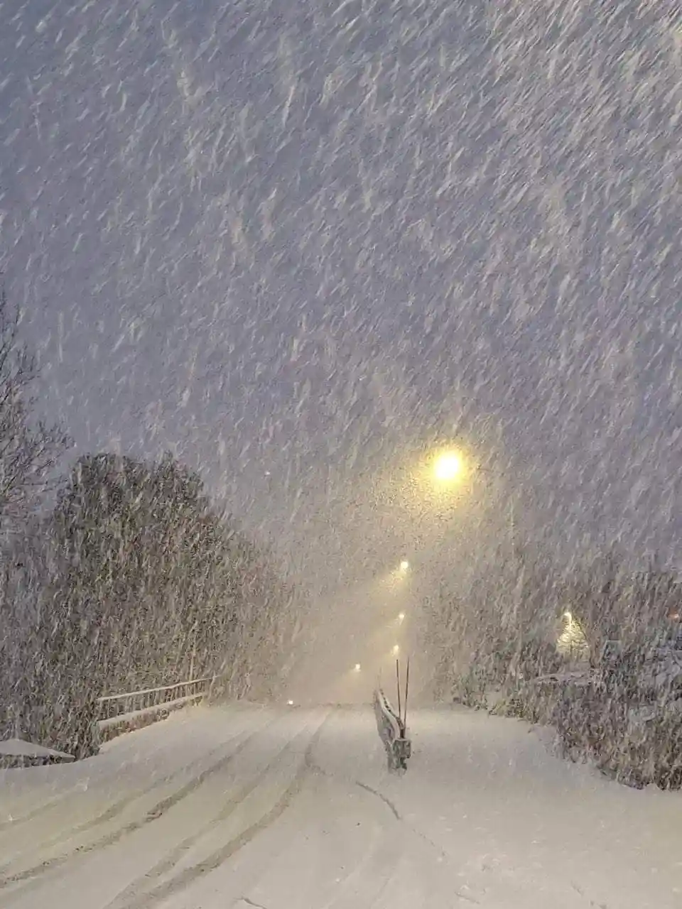 A nighttime photo of a snow-covered road with heavy snowfall obscuring visibility, streetlights glowing faintly through the…