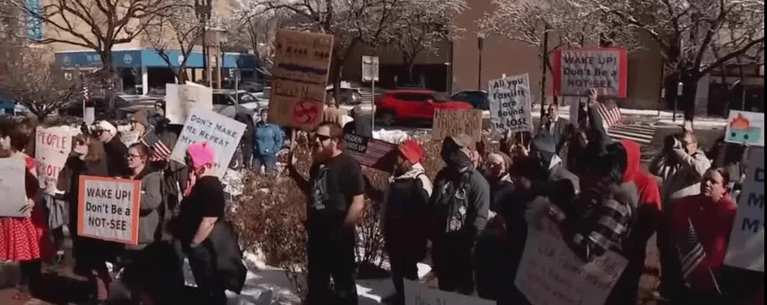 A crowd of protesters holding various handmade signs outdoors in winter, with snow on the ground and bare trees. Several…