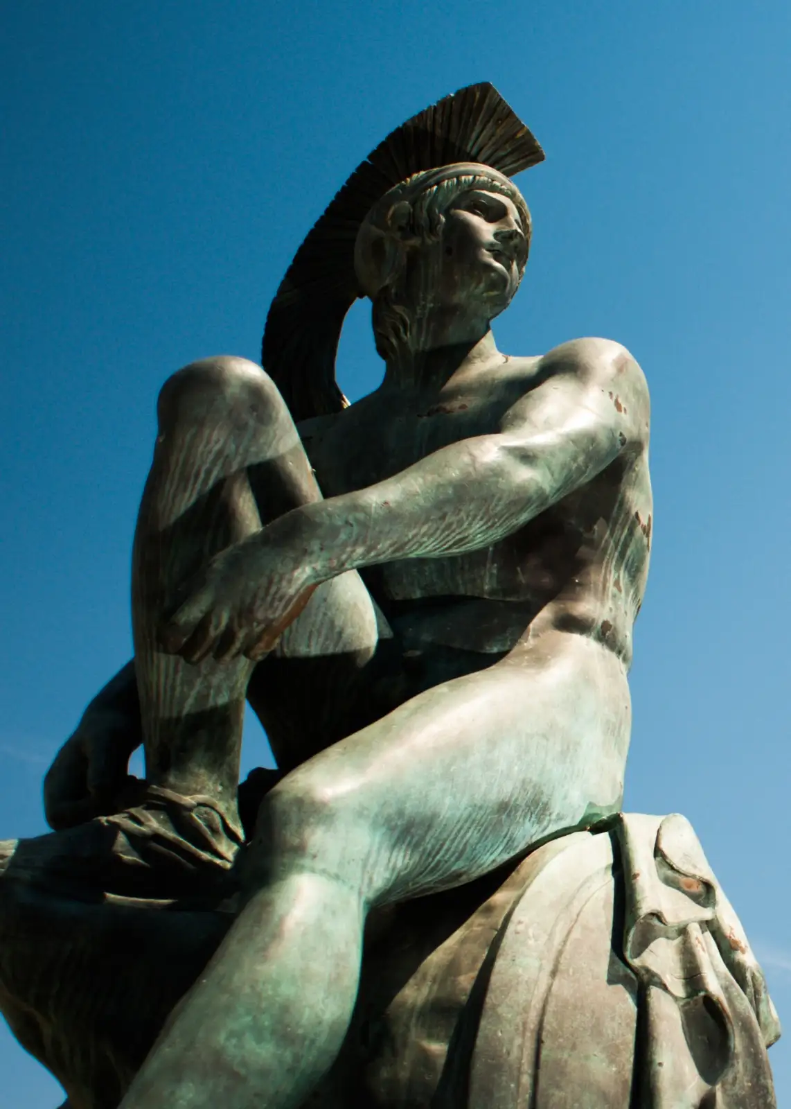 A bronze statue of a muscular, helmeted classical warrior sits cross-legged against a clear blue sky, looking upward with a…
