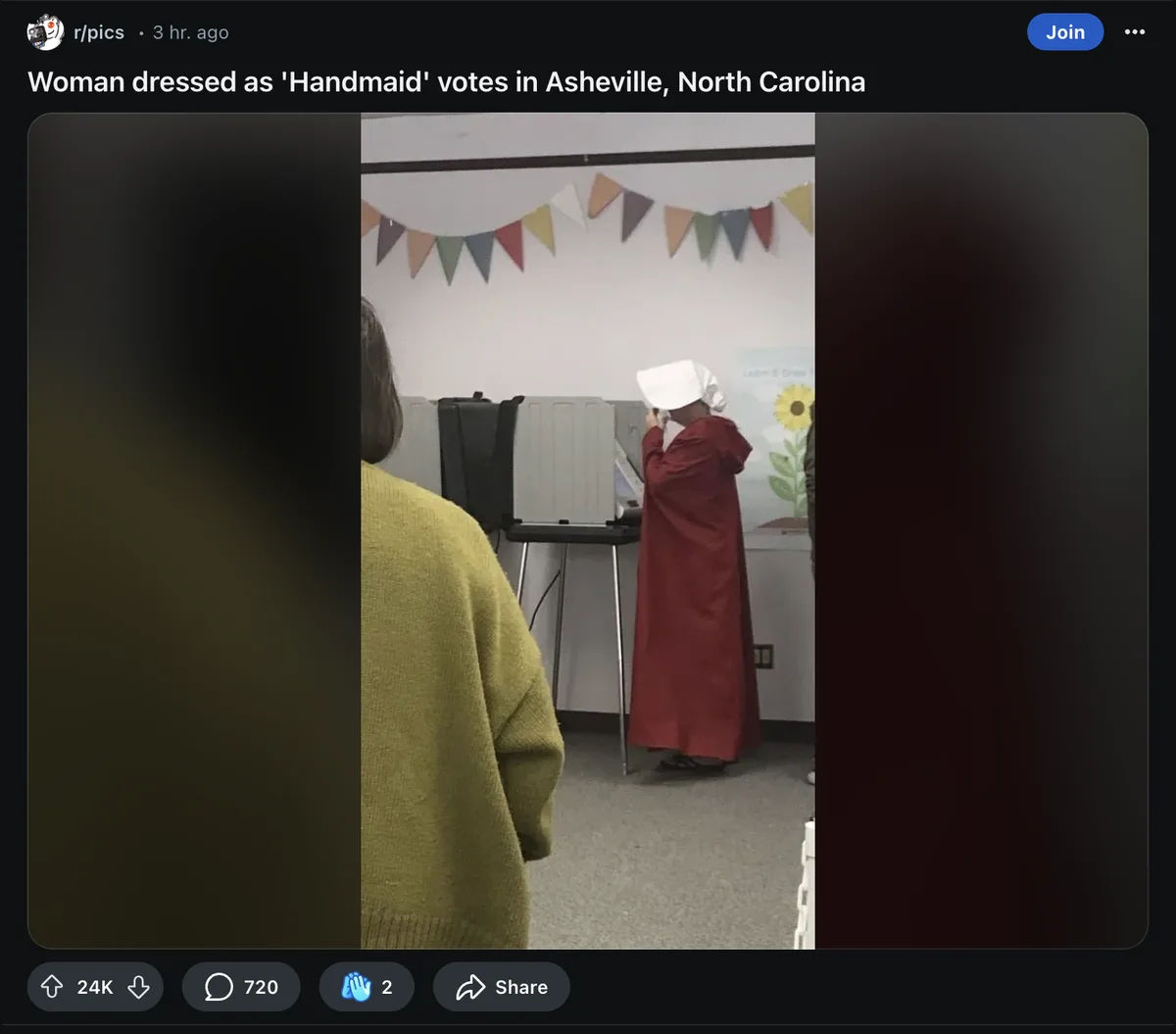 A person wearing a red robe and white bonnet, resembling a Handmaid from 'The Handmaid's Tale,' is standing at a voting…