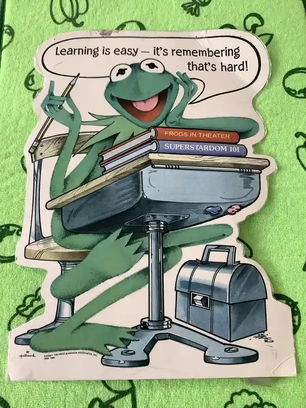 A cartoon illustration of Kermit the Frog sitting at a school desk, holding a pencil and smiling while looking at the…