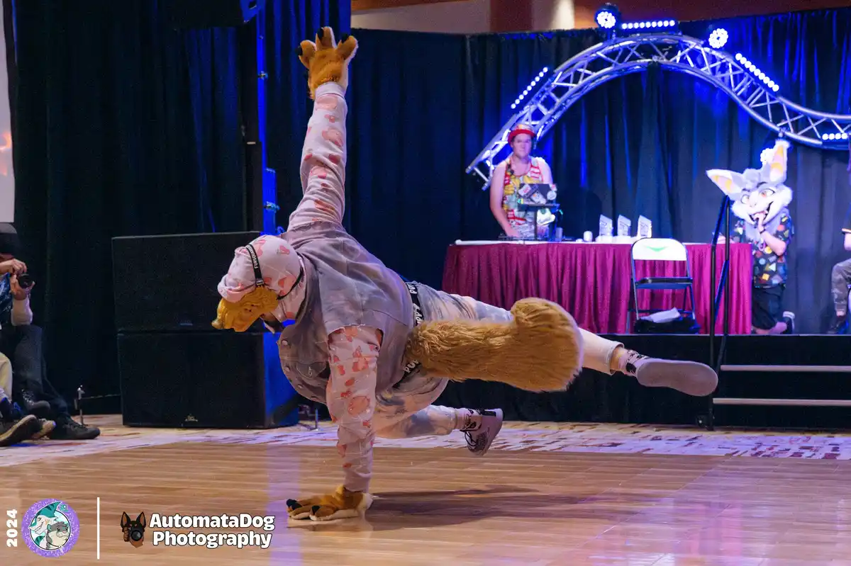 A person in a full-body furry costume, resembling a fox or dog, performs a dynamic breakdancing move on a wooden stage…