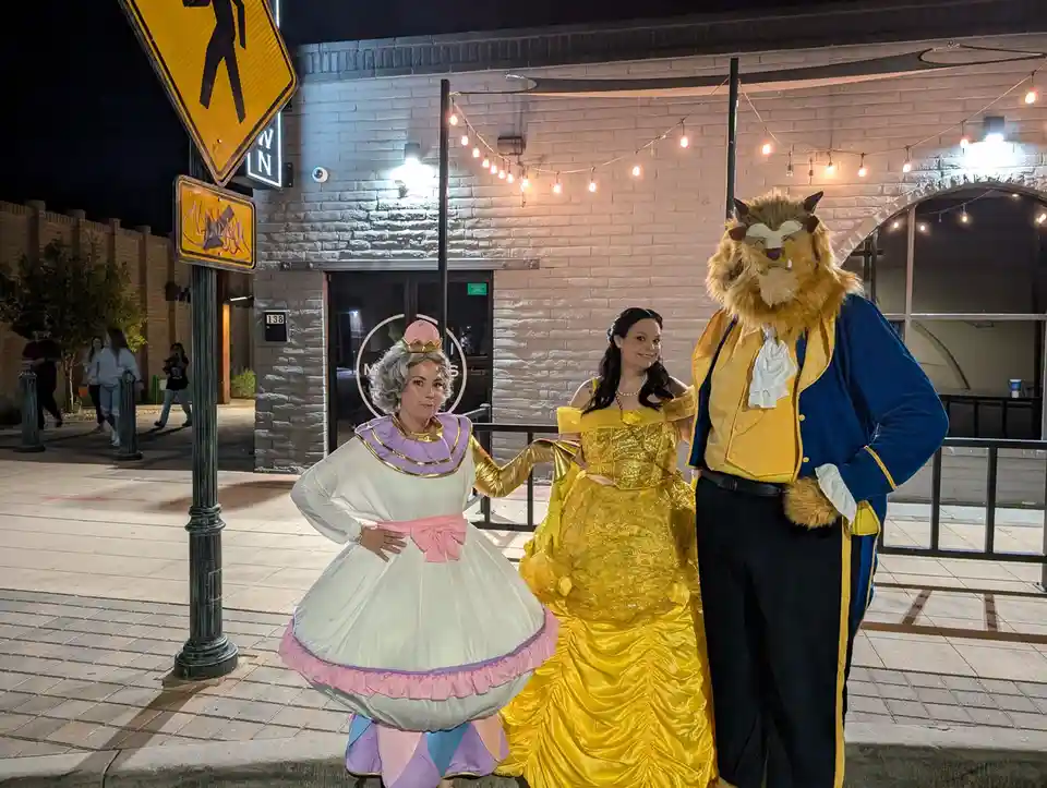 Three people dressed as characters from Beauty and the Beast stand on a sidewalk at night: a woman in a white and pink dress…