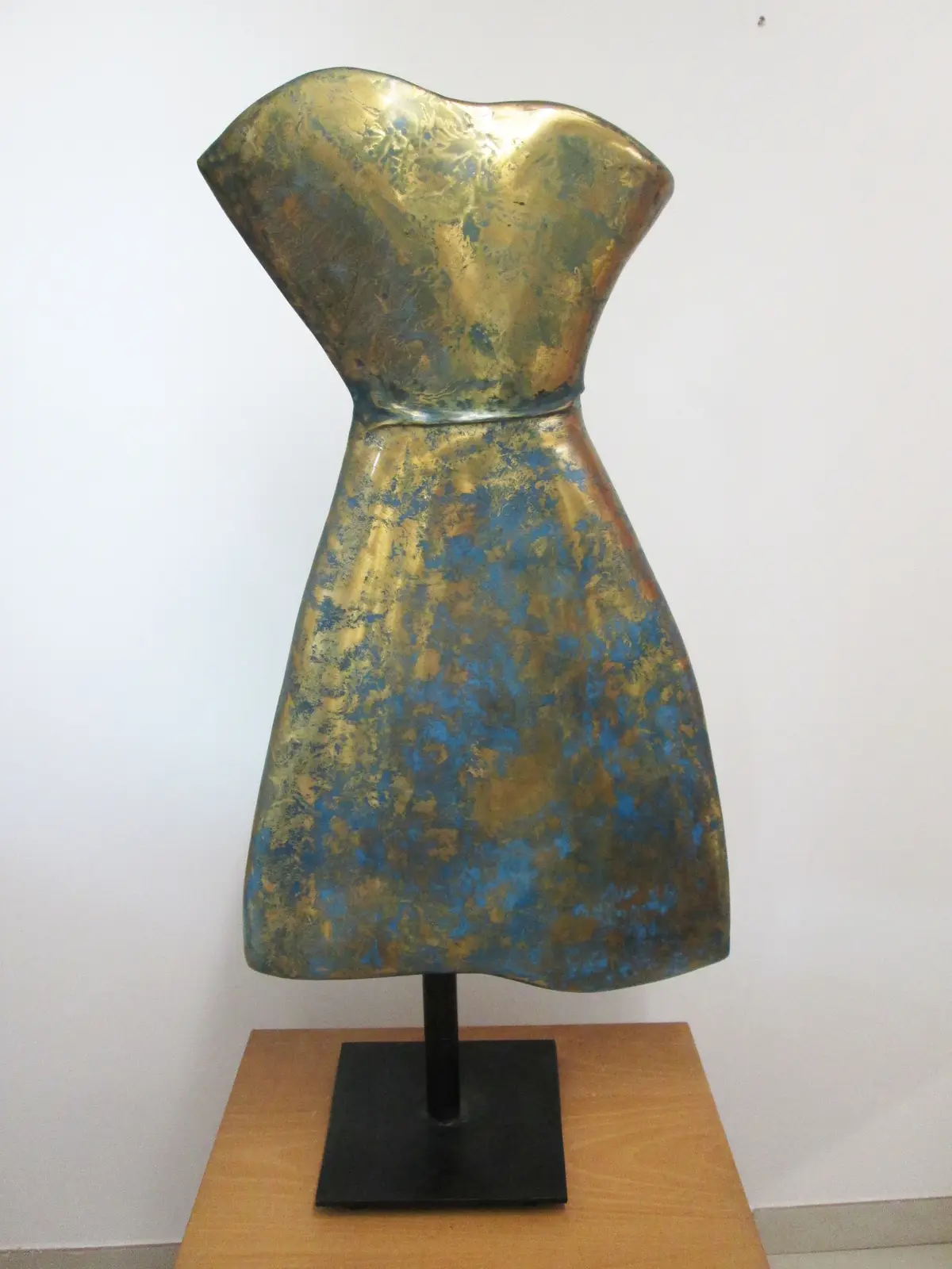 A metallic sculpture shaped like a dress with gold and blue patina stands on a black pedestal atop a wooden table against a…