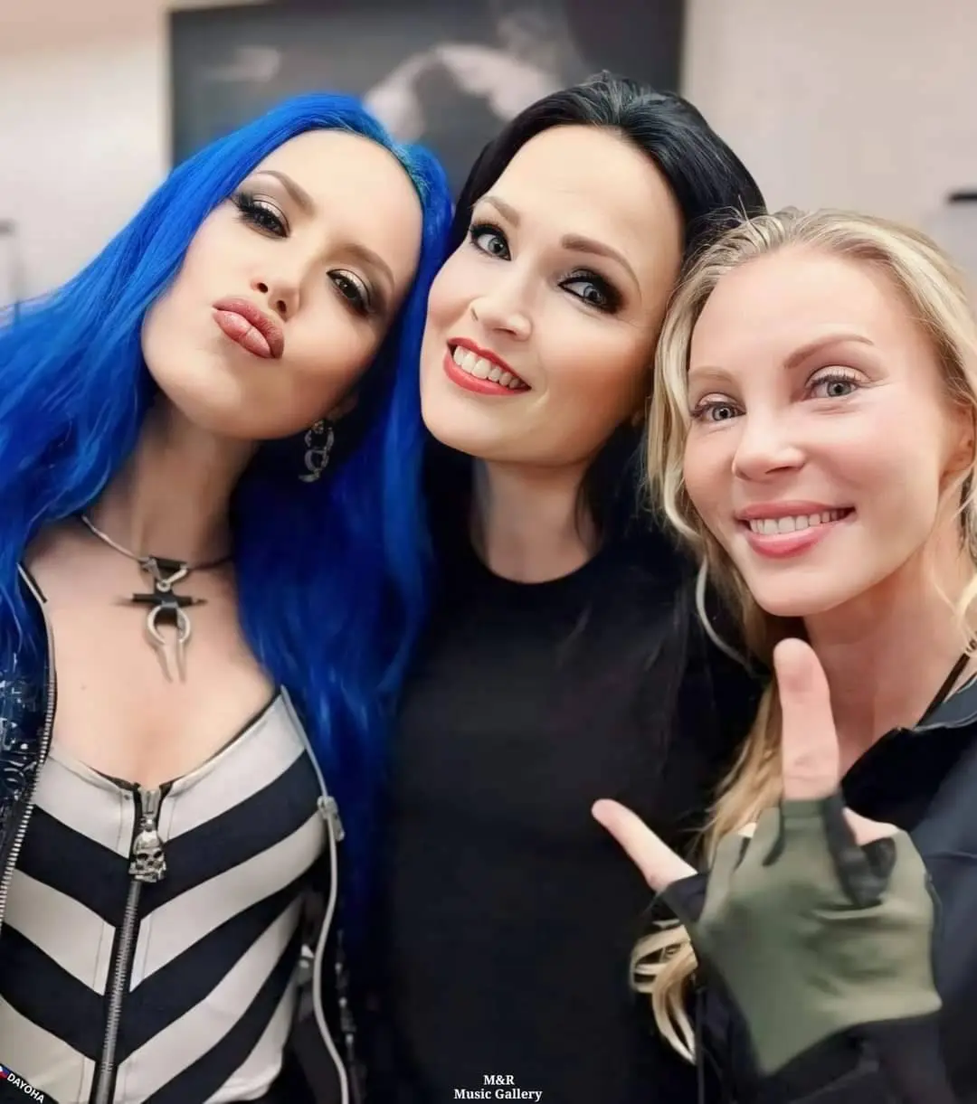 Three women pose closely for a selfie; the woman on the left with bright blue hair makes a pouty face, the center woman…
