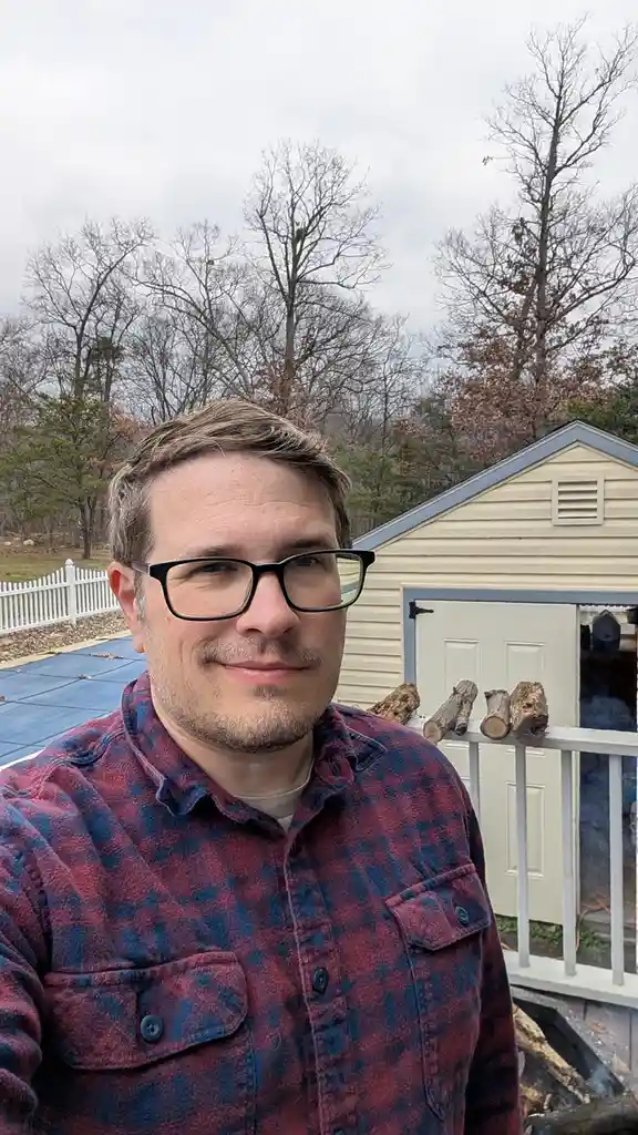 A man with glasses and a plaid shirt smiles at the camera outdoors, standing in front of a shed with logs on its railing…
