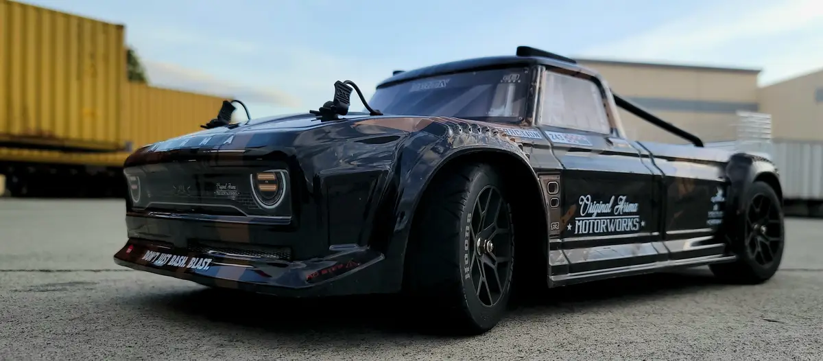 A black remote-controlled truck with racing decals and 'Original Arma MOTORWORKS' branding is parked on asphalt. The front…