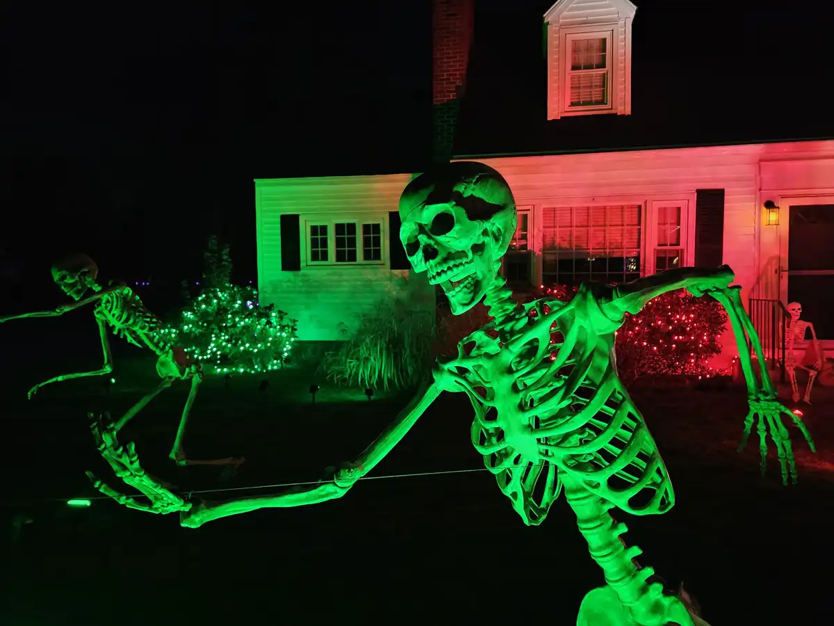 A large, green-lit Halloween skeleton is posed in a yard at night, holding a taut rope with both hands as if pulling…