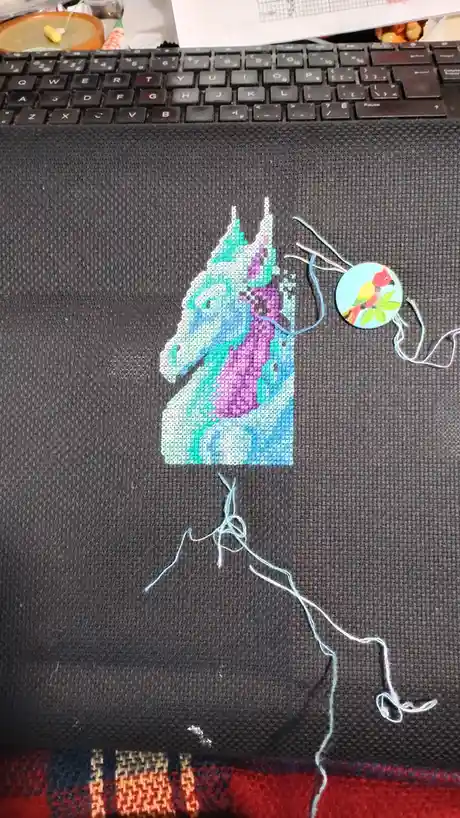 A partially completed cross-stitch project of a colorful unicorn head on dark fabric, with loose threads dangling below and…