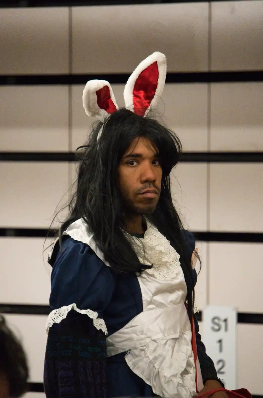 A person with long dark hair and a beard wears a blue and white maid-style costume with white lace trim and large…