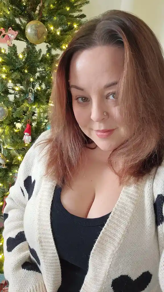 A woman with shoulder-length brown hair and a nose piercing poses in front of a decorated Christmas tree with lights and…