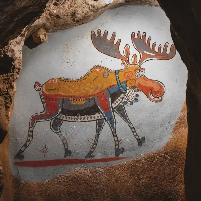 A stylized, cartoonish moose painting on a cave wall, decorated with colorful patterns and jewelry, with overlaid text…