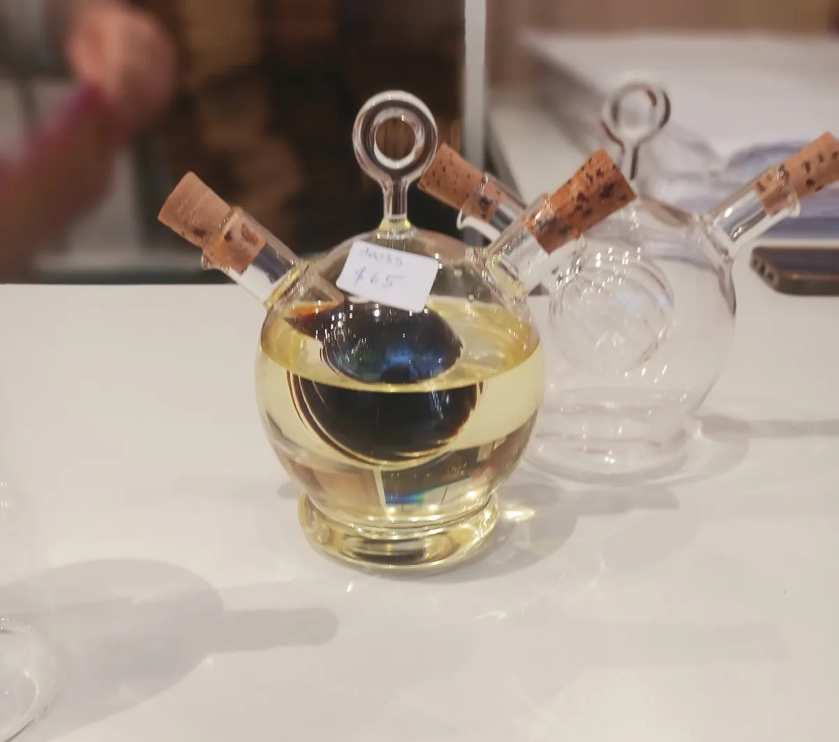 A clear glass decanter filled with a golden liquid sits on a white table, with a handwritten price tag reading 'Auss $65'…