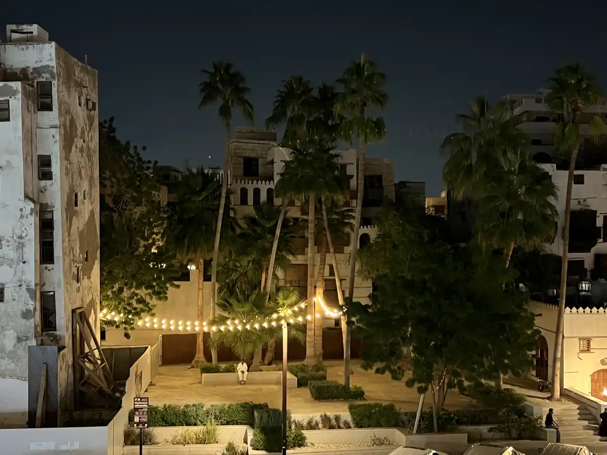 A nighttime photo of a courtyard with palm trees, string lights, and buildings, featuring a person sitting alone on a bench…