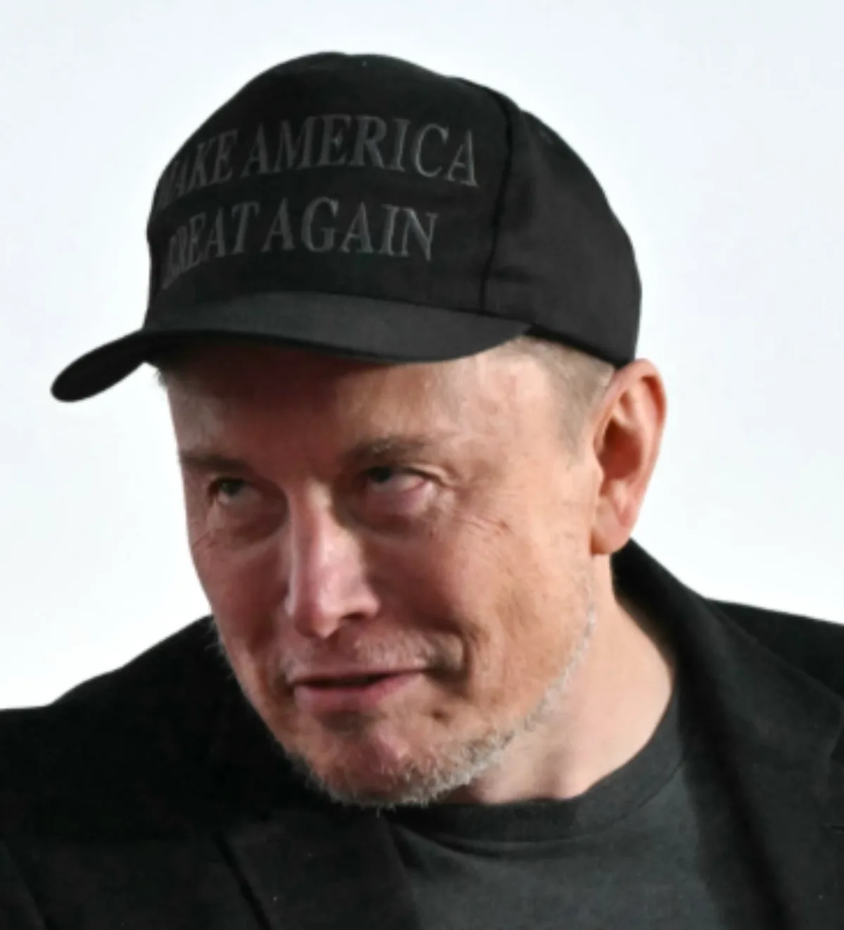 A man with short graying hair and a goatee wears a black baseball cap with the text 'MAKE AMERICA GREAT AGAIN' in gray…