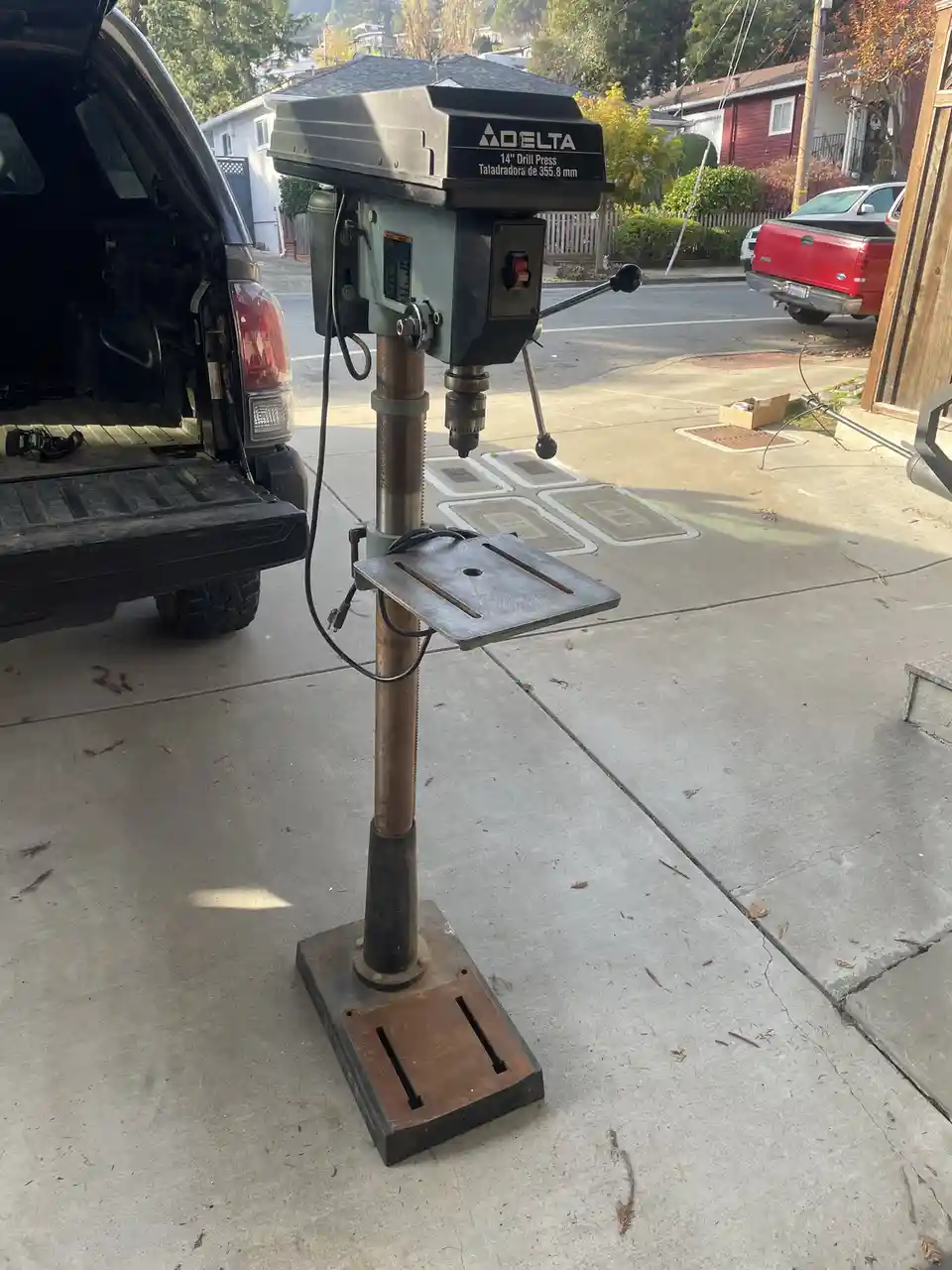 A Delta 14-inch drill press stands on a concrete driveway next to an open black SUV, with a red pickup truck and suburban…